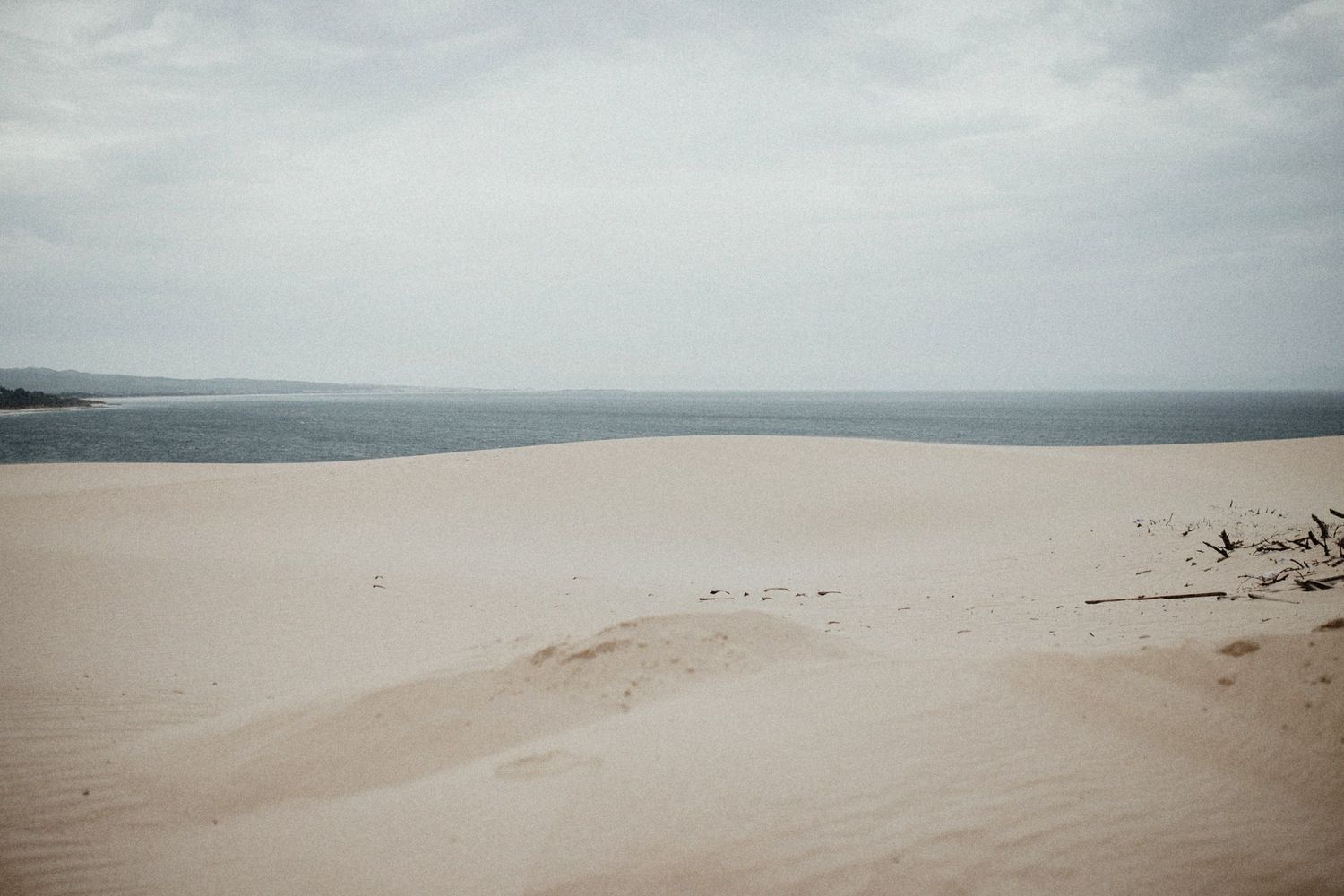 Expansive sandy dunes meet the ocean under overcast skies in moody landscape shot.