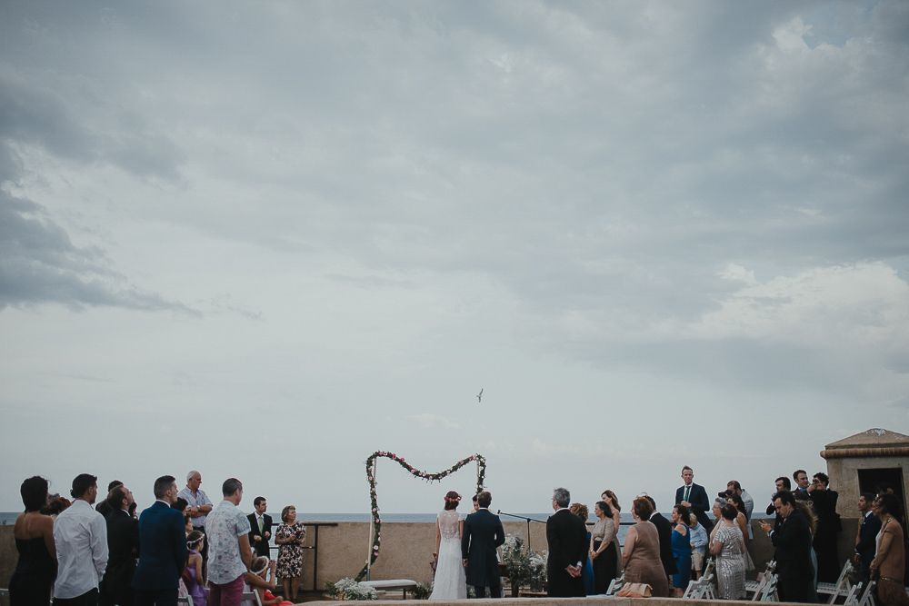Wedding guests gather outdoors under cloudy skies for a ceremony by the sea.