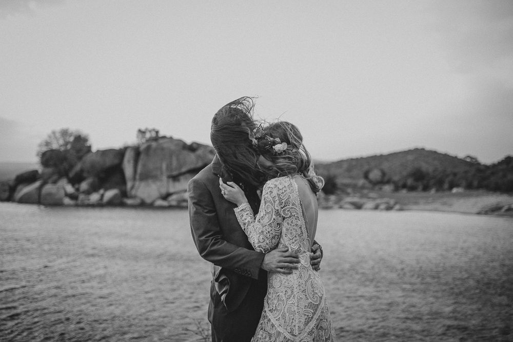 Black and white photo of a tender embrace by the water with boulders and hills in the distance.
