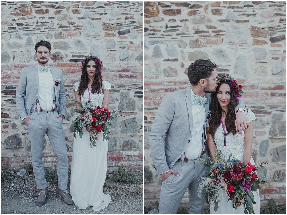 A couple poses against a rustic stone wall backdrop, with the bride holding a vibrant red bouquet.