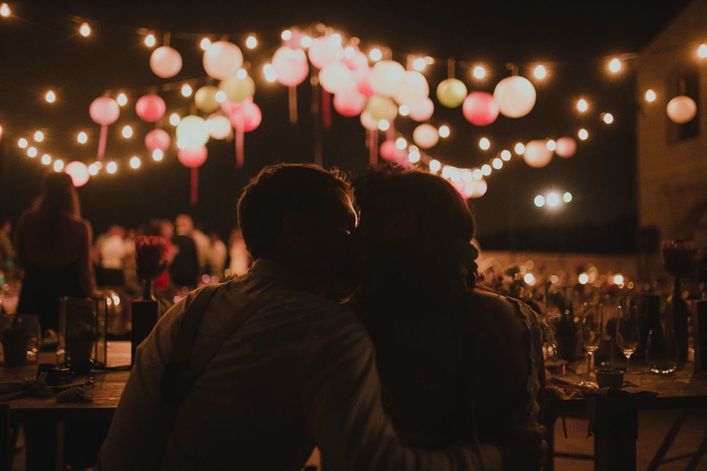 Couple enjoys romantic moment under string lights and pink lanterns at outdoor evening event.
