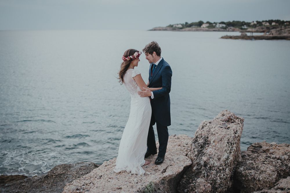 Romantic wedding portrait of couple standing on coastal rocks with misty ocean backdrop.
