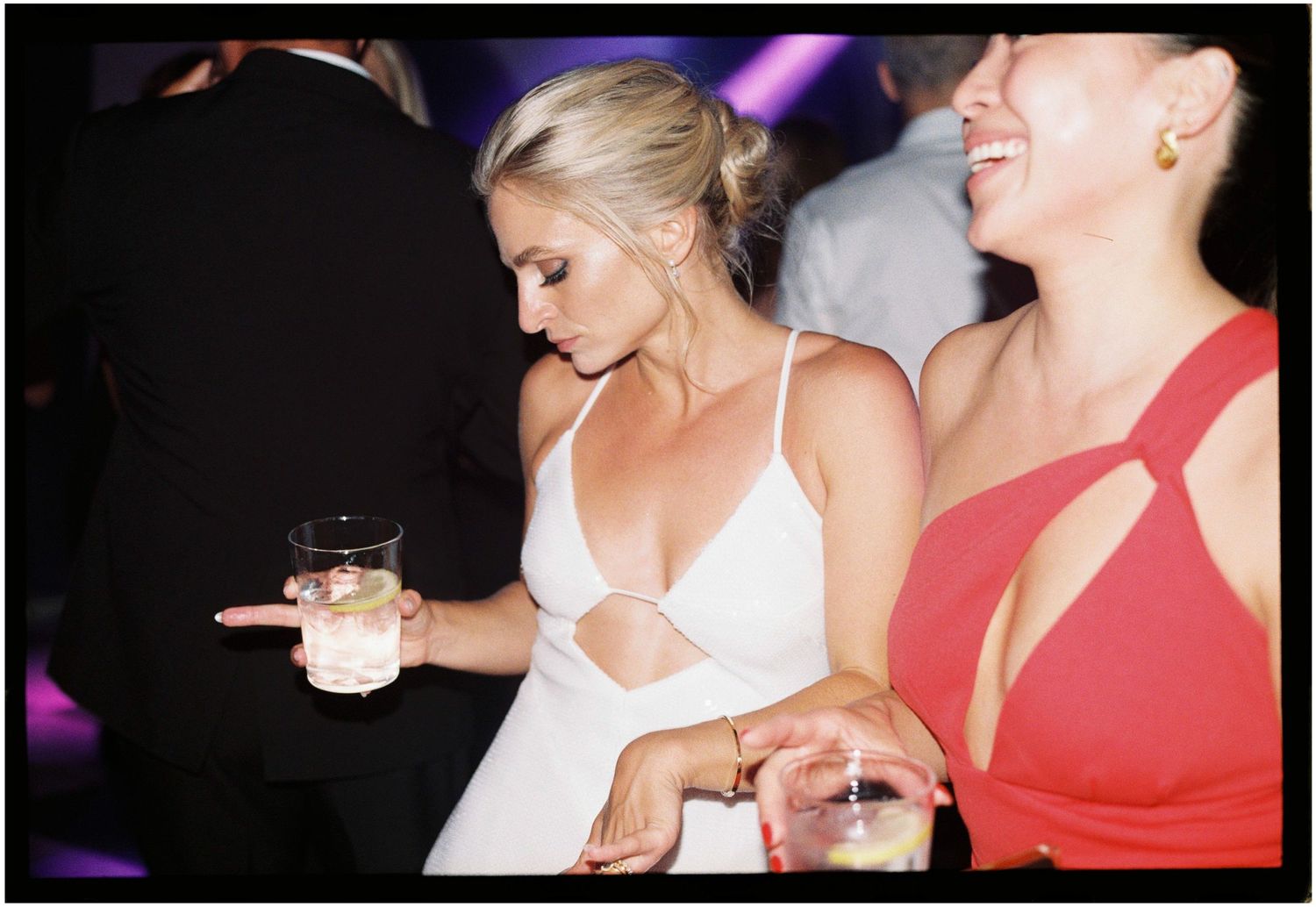 Two people celebrating with drinks at a party, with one wearing a white dress and another in red.