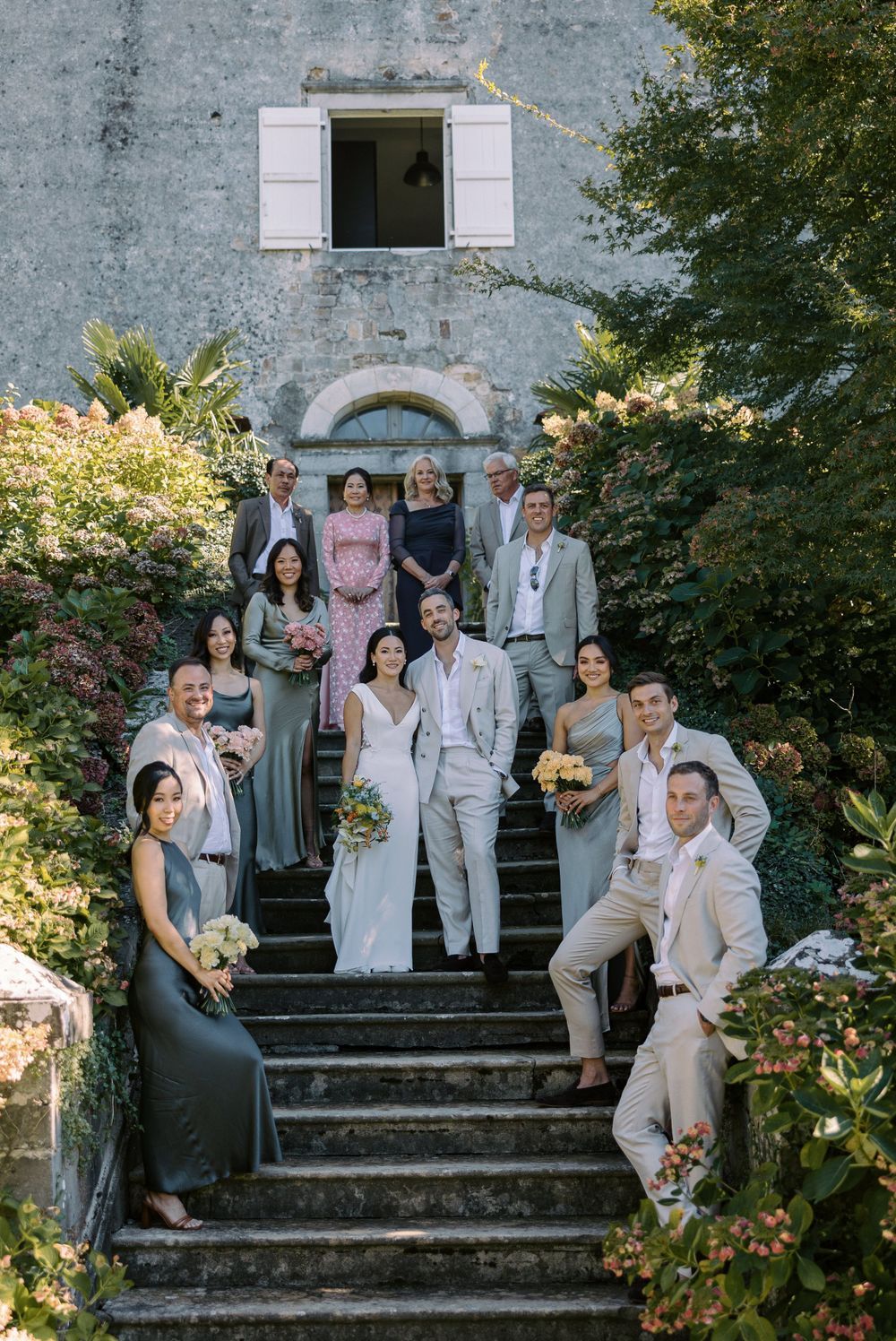 Wedding party poses on stone steps outside historic building with white shuttered windows.