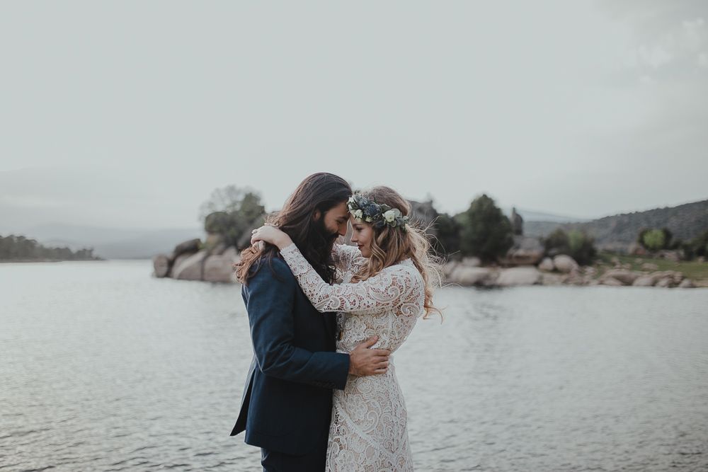 Romantic wedding couple embraces on rocky shoreline with mountains and lake in background.