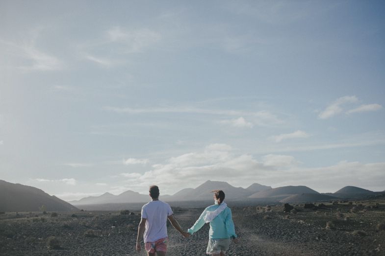 Two figures hold hands while standing in a vast desert landscape with mountains in the background.