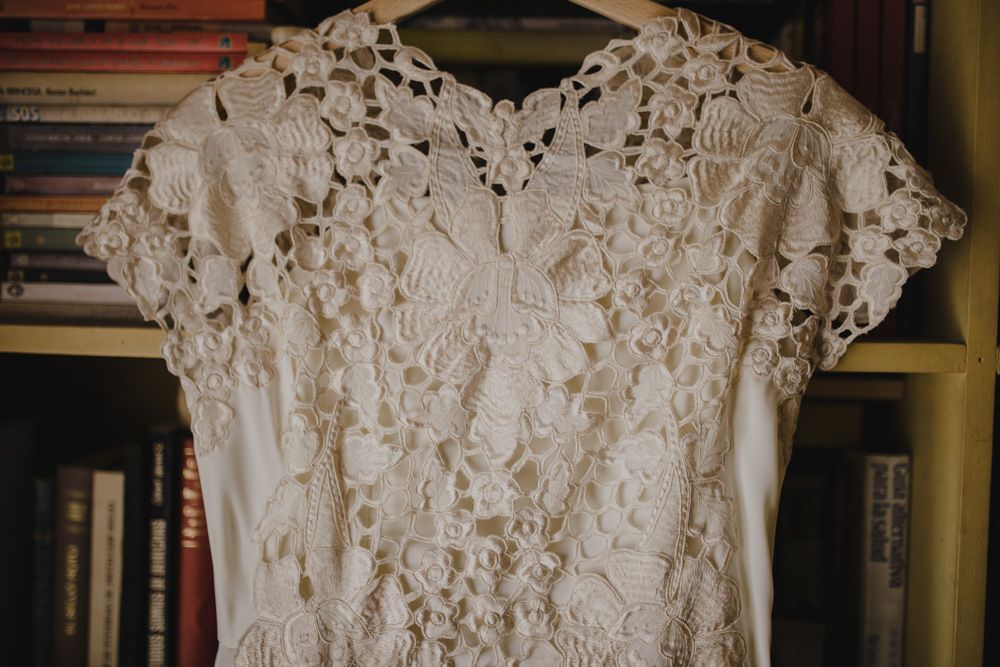 Delicate lace wedding dress with intricate floral patterns and cap sleeves hanging against a bookshelf.
