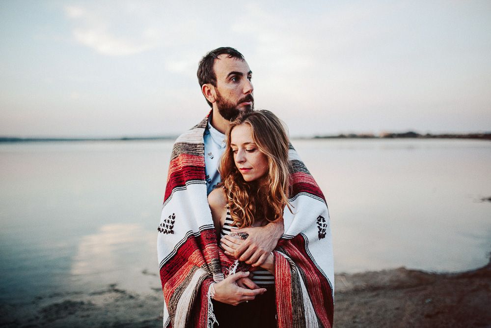 Couple embracing while wrapped in patterned blanket against tranquil water backdrop.