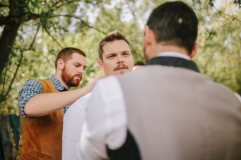 Wedding party members assist with formal wear adjustments in natural outdoor setting.