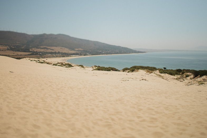 Scenic coastal landscape with sandy dunes overlooking a blue bay and mountain range.