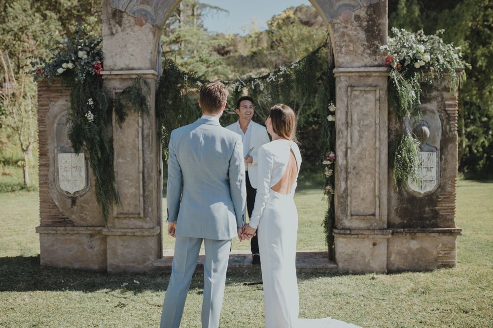 A couple stands under an ornate stone archway decorated with greenery during their wedding ceremony.