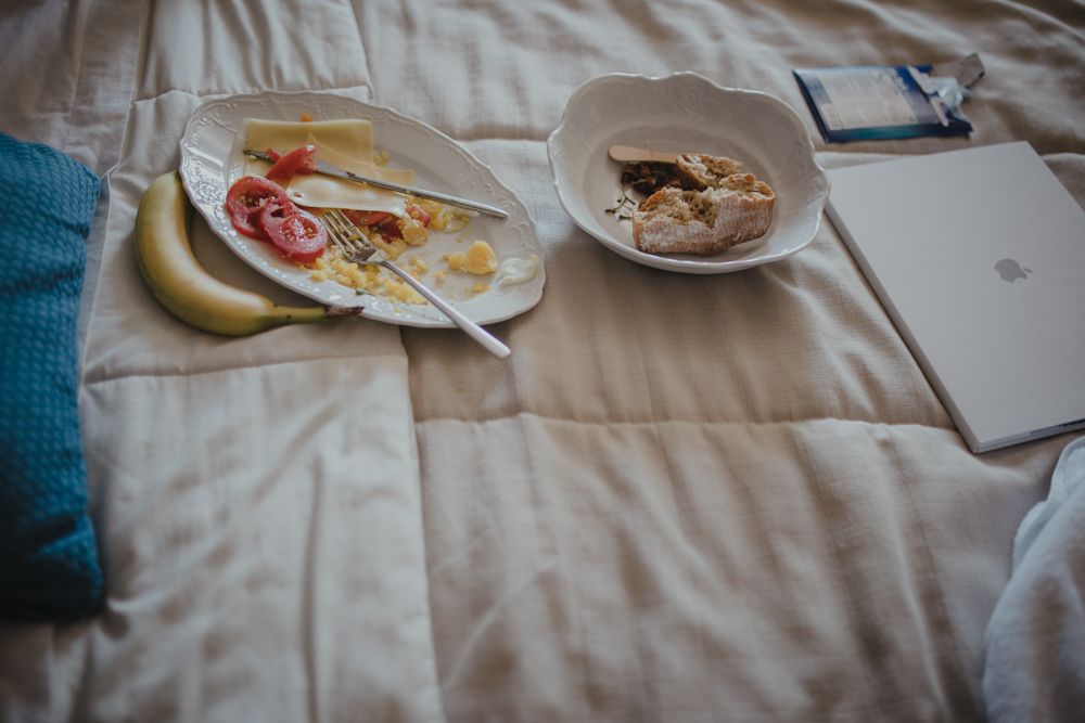 Breakfast in bed with plates of food and a laptop on white bedding.