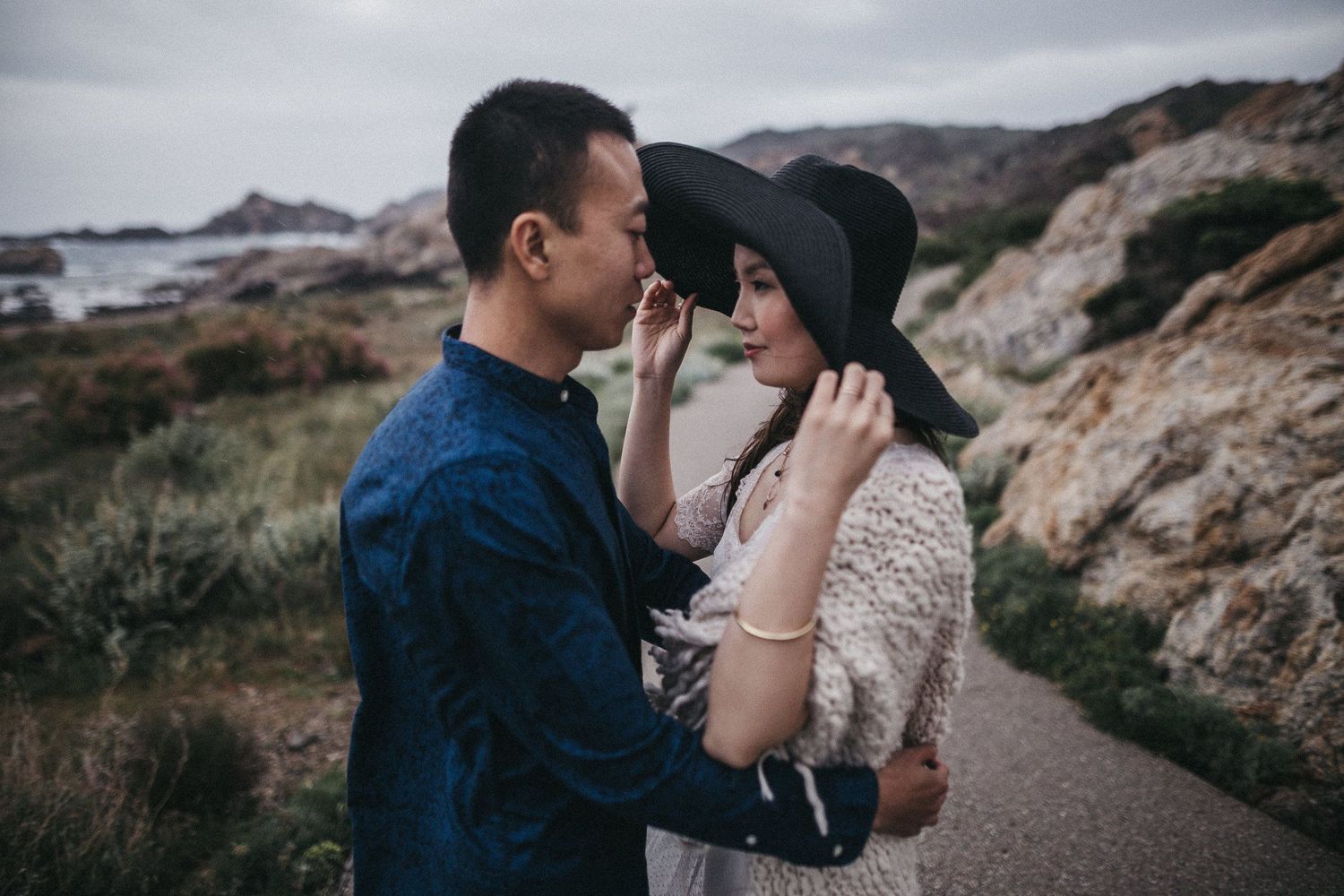 Intimate moment shared on a coastal path with rocky cliffs and ocean backdrop.