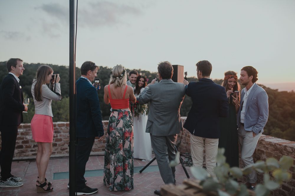 A group of people gather for an outdoor event at sunset on a balcony or terrace overlooking scenic views.