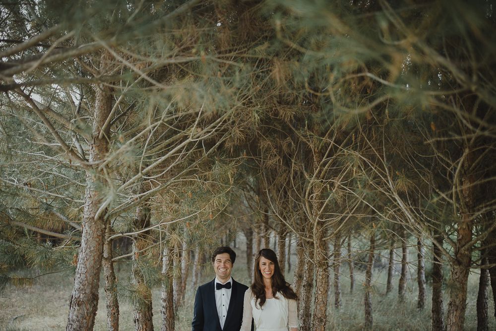 Newlyweds walk through a misty field with evergreen trees in the background.