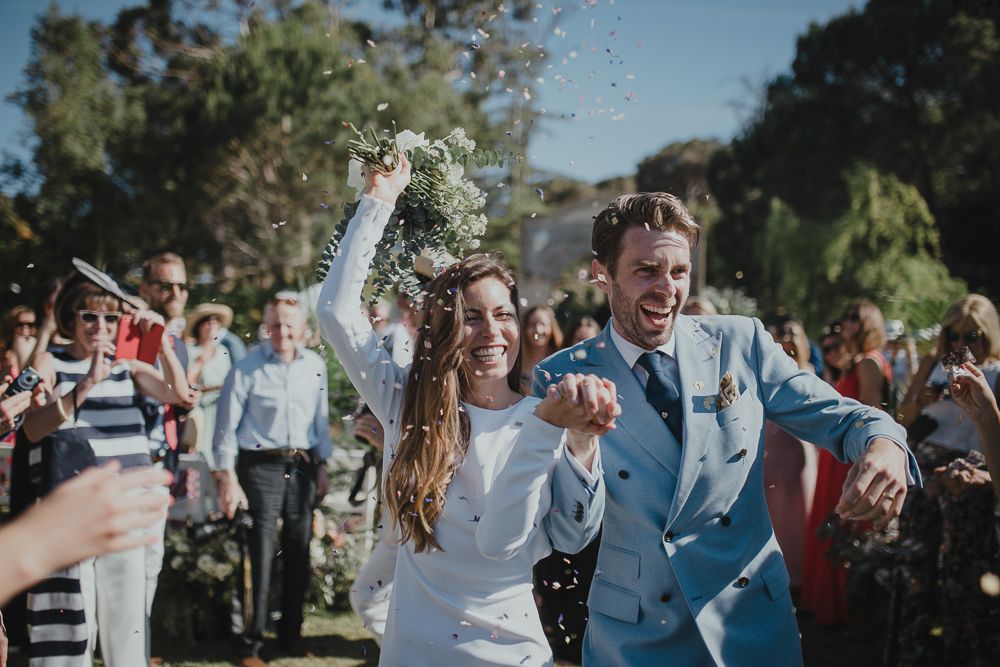 Joyful newlyweds celebrate during outdoor ceremony as guests shower them with confetti under sunny skies.