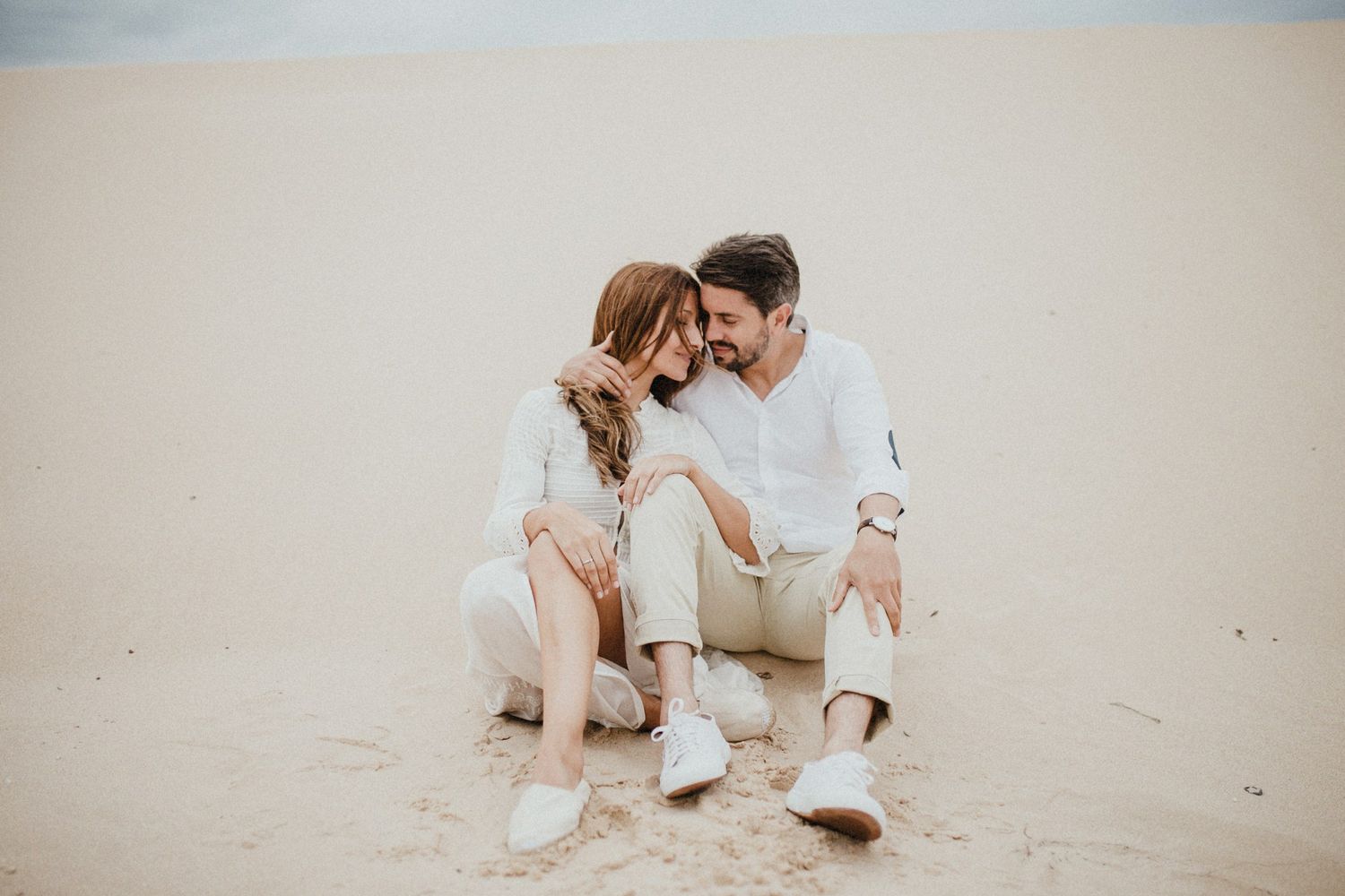 Couple in light colored clothing sitting close together on sandy beach during overcast day.