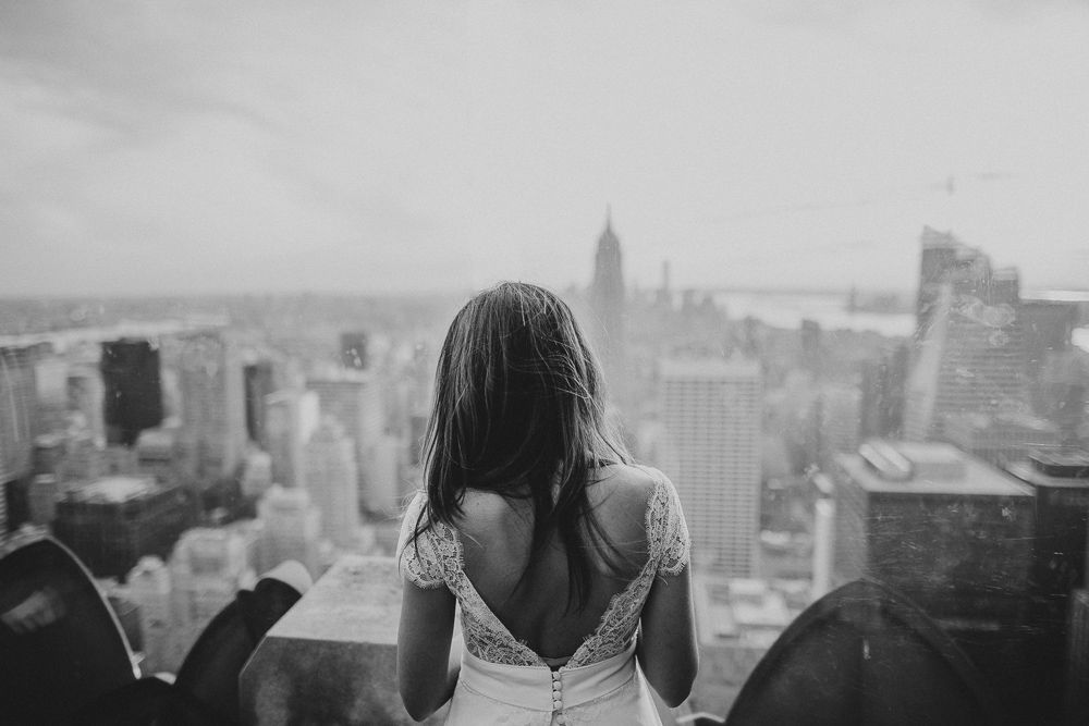 A backlit figure in a white dress looks out over the Manhattan skyline from a high vantage point.