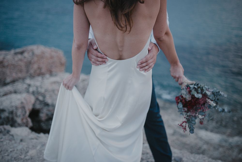 Back view of flowing white wedding dress against dramatic coastal background.