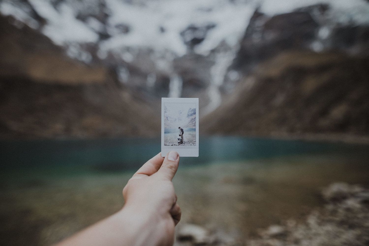 A hand holding up a photograph against a backdrop of mountains and a turquoise alpine lake.