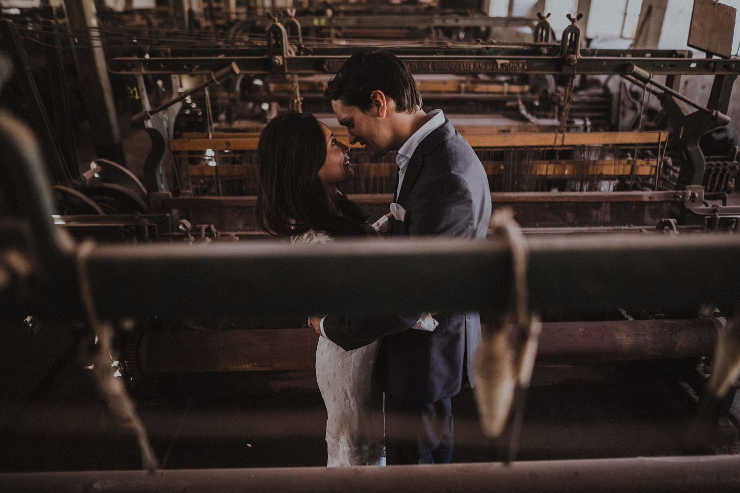 Romantic couple embraces between vintage industrial machinery in a rustic warehouse setting.