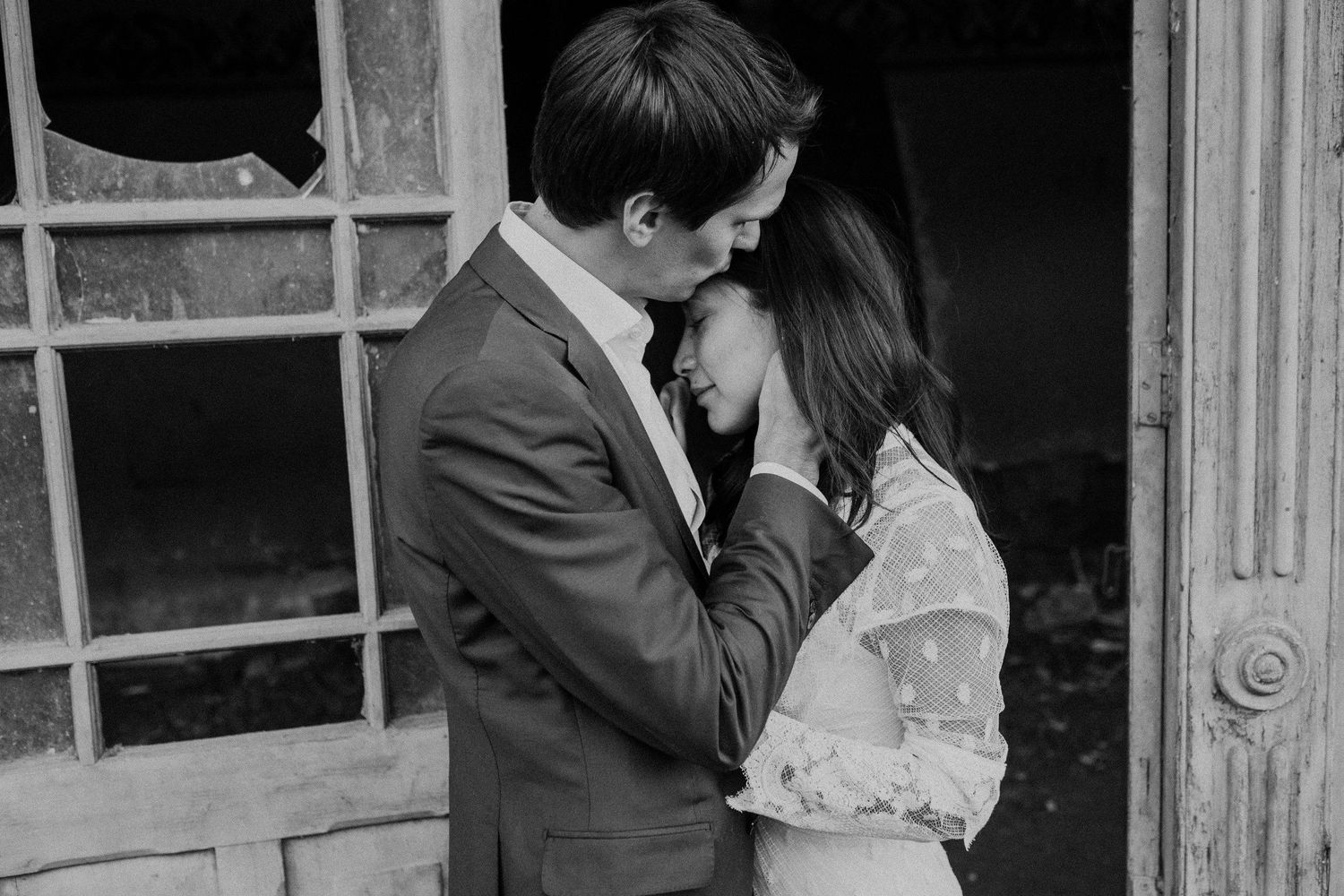 Black and white portrait of a tender moment between a couple in wedding attire by a weathered doorway.