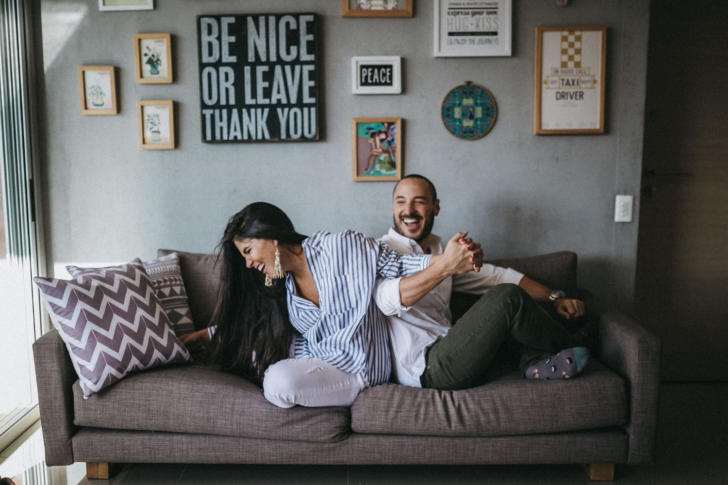 A playful couple relaxes on a gray couch beneath wall art reading 'Be Nice or Leave'.