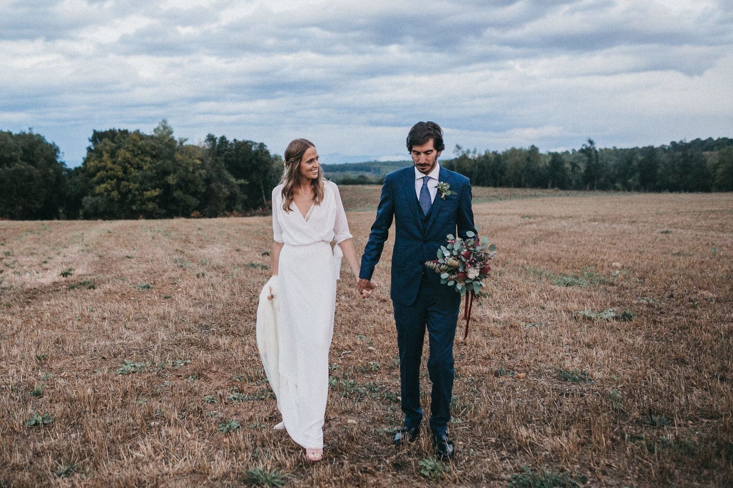 Newlyweds walking hand in hand through an open field under cloudy skies.