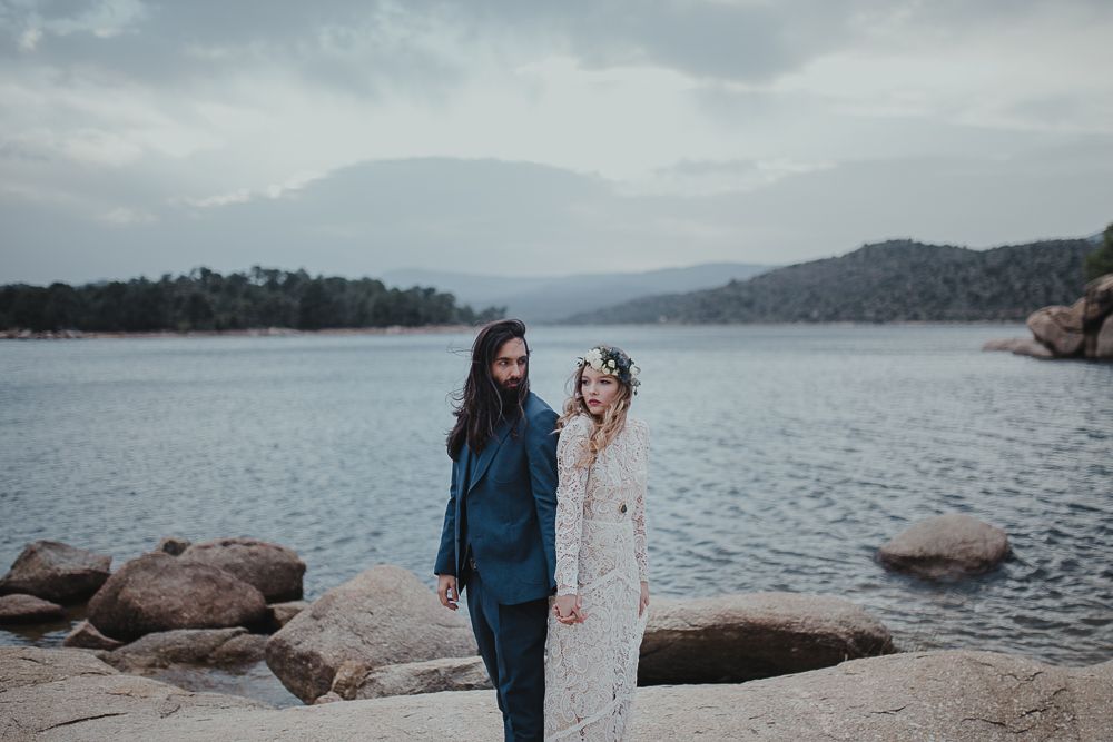 A couple stands together on rocky terrain beside a peaceful mountain lake.