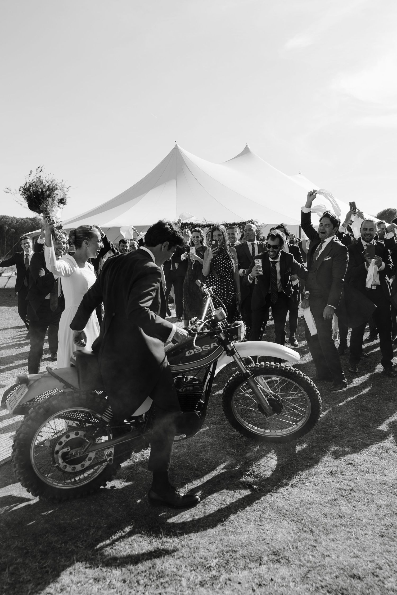 Black and white photo of wedding guests cheering as someone rides a motorcycle near outdoor event tents.