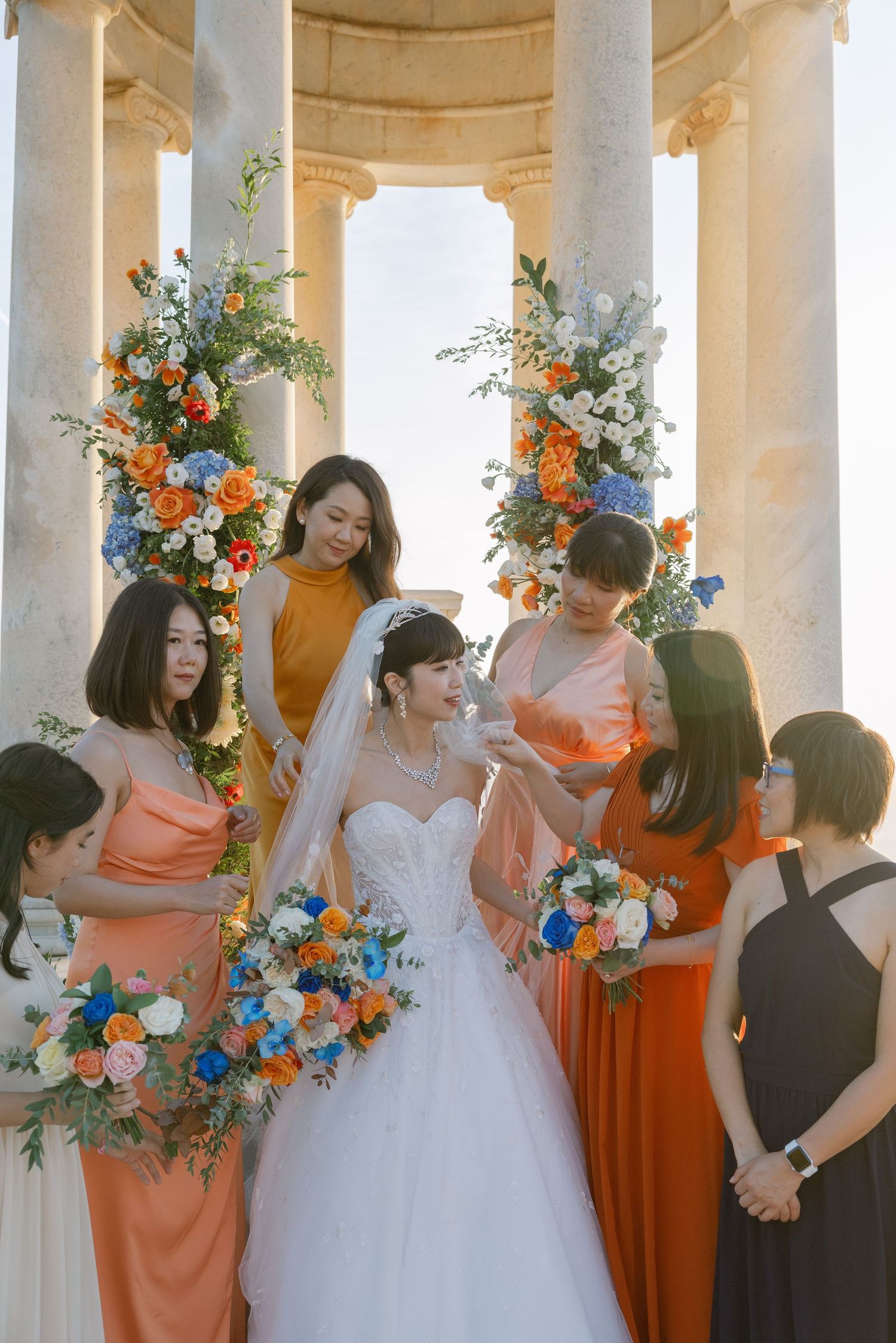 Bride in white gown with bridesmaids in coral dresses holding colorful bouquets at an elegant column wedding venue.