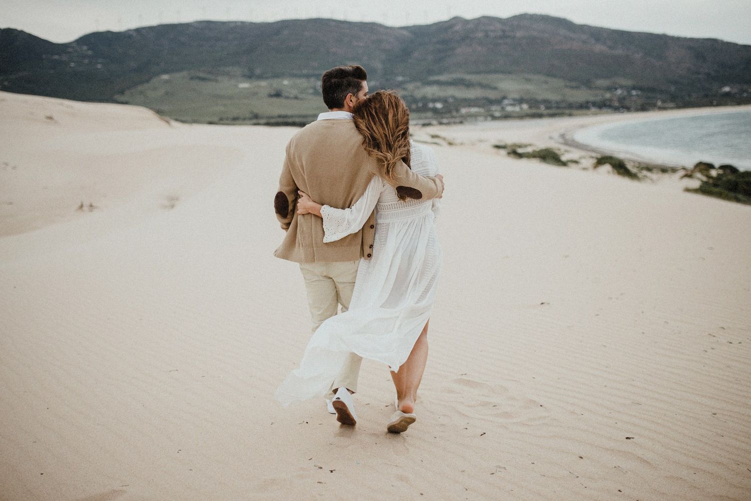 Couple walking together across sweeping sand dunes with mountains in background.