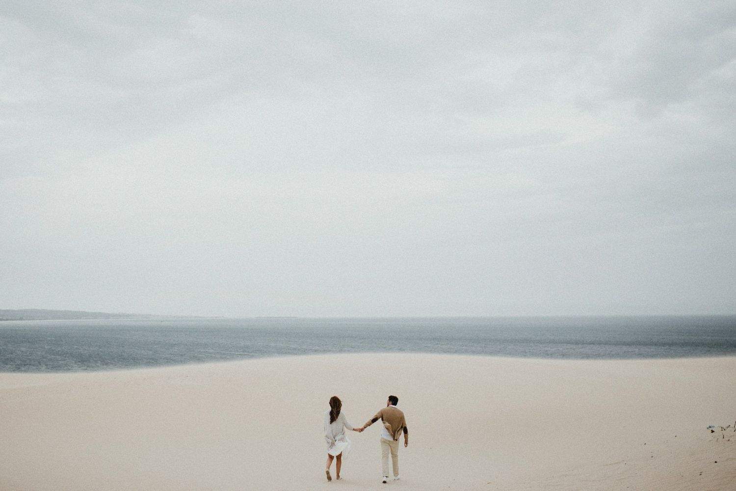 Two figures walking together on expansive sand dunes against cloudy horizon.