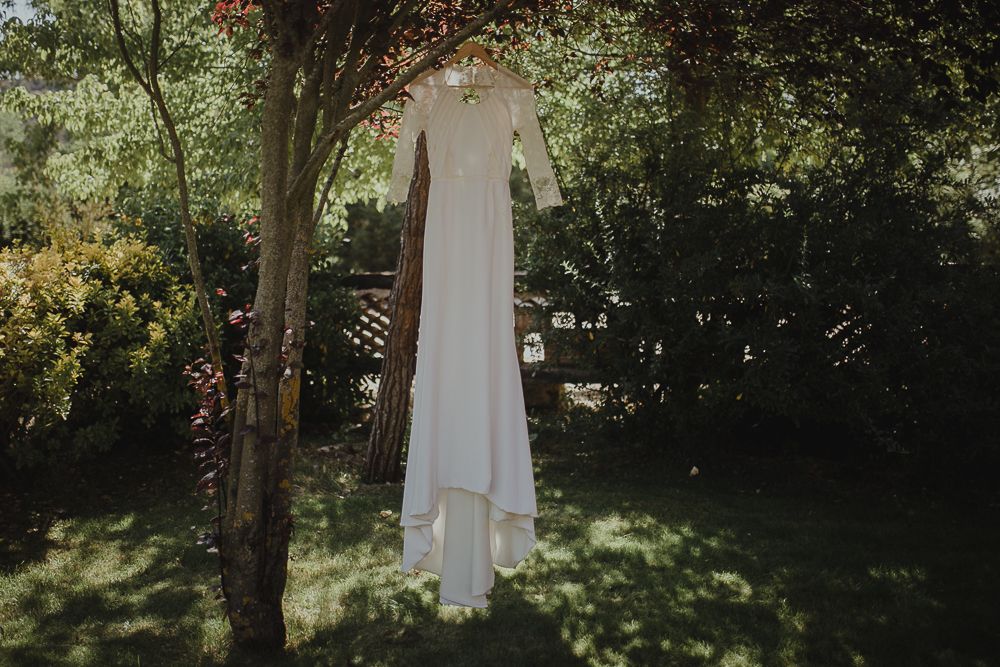 White wedding dress hanging from tree branch in sunlit garden setting.