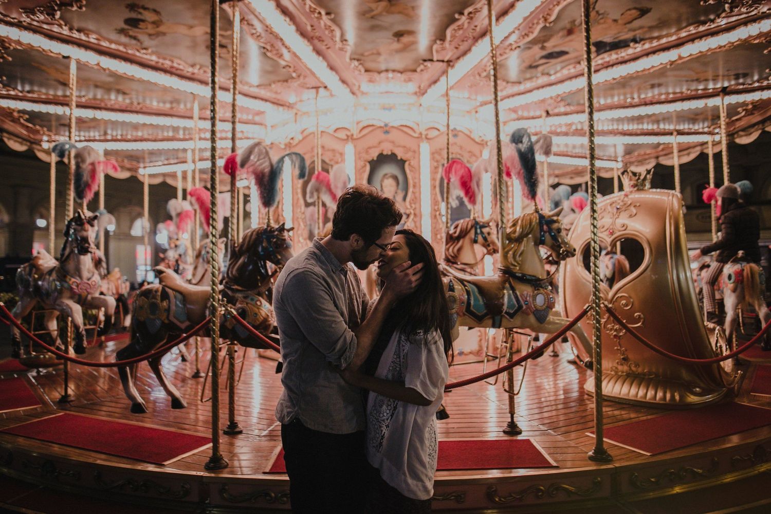 A romantic couple embracing near an illuminated carousel at night.