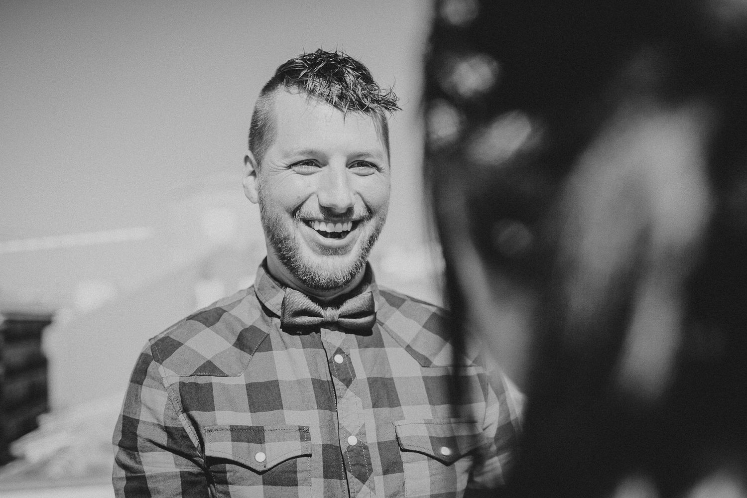 A smiling person in a red plaid shirt poses for a black and white portrait outdoors.