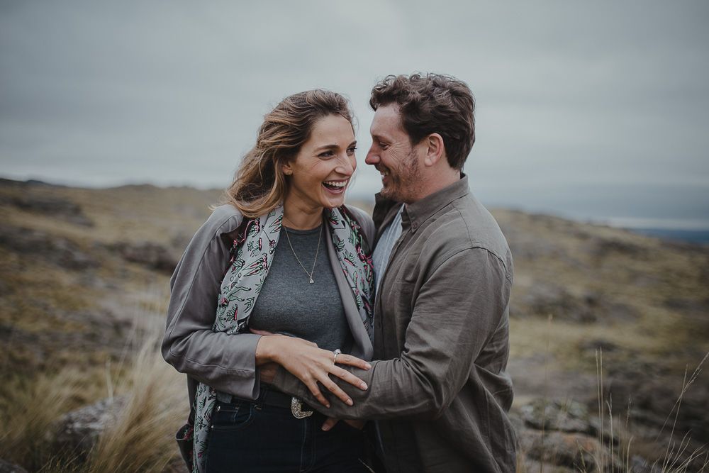 A couple laughs together during an intimate moment on a windy hilltop.