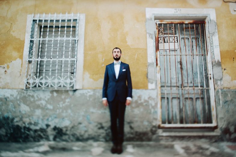 Figure stands against weathered wall with barred window and rustic door in formal attire.