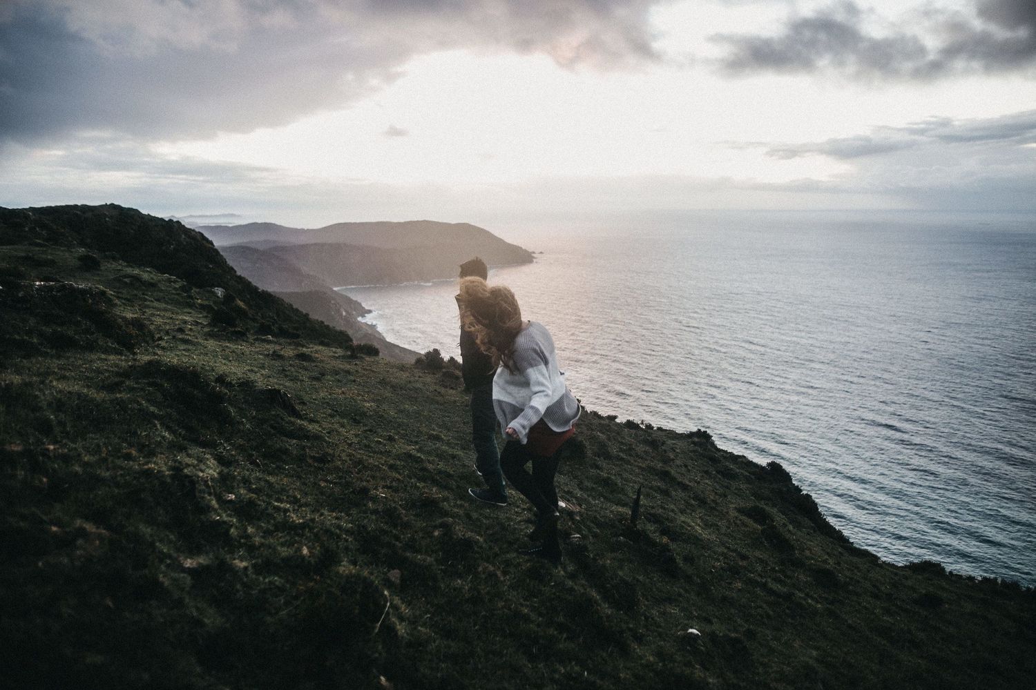 A couple stands on a cliff overlooking a dramatic coastal sunset view.