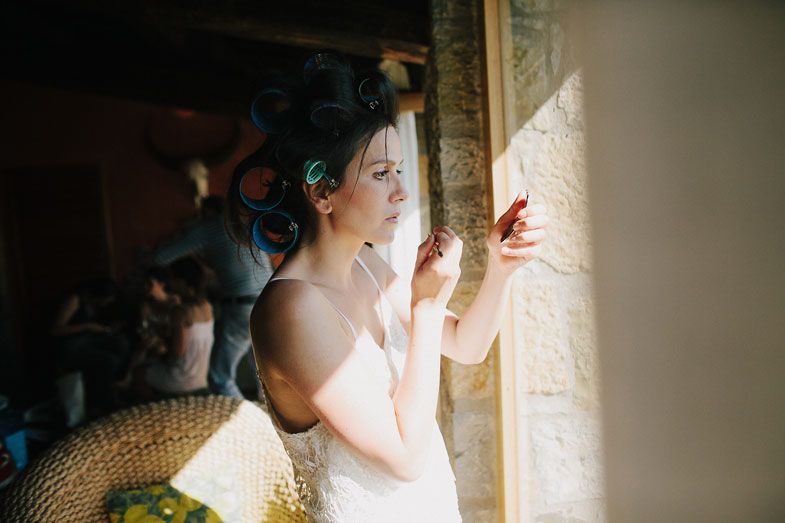 A bride in white peeks through a curtain with gentle sunlight streaming in during wedding preparations.