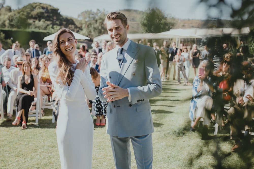 A couple laughing together on their wedding day with guests visible in the background.