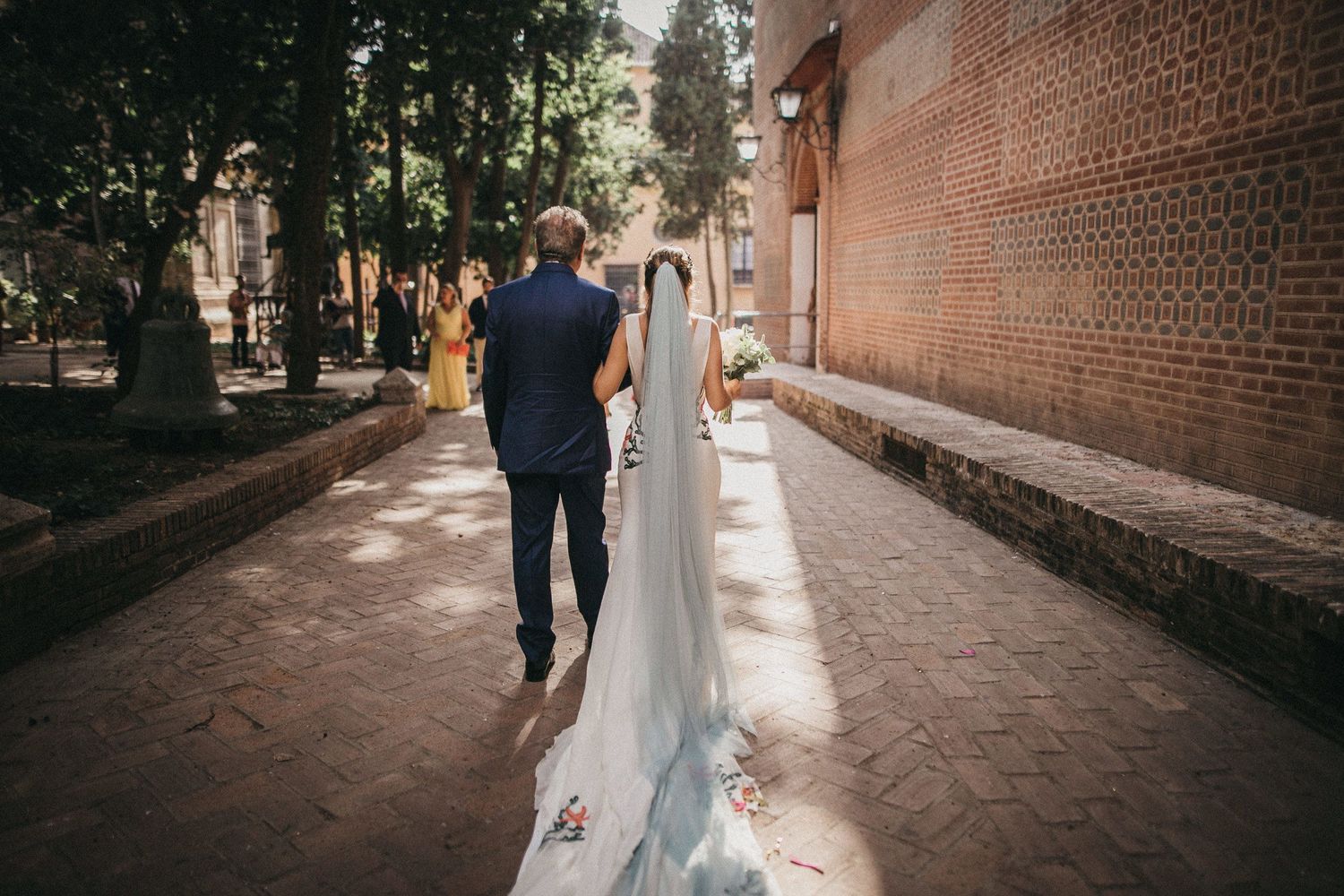 A wedding couple walks down a brick-lined path with dappled sunlight streaming through the trees.