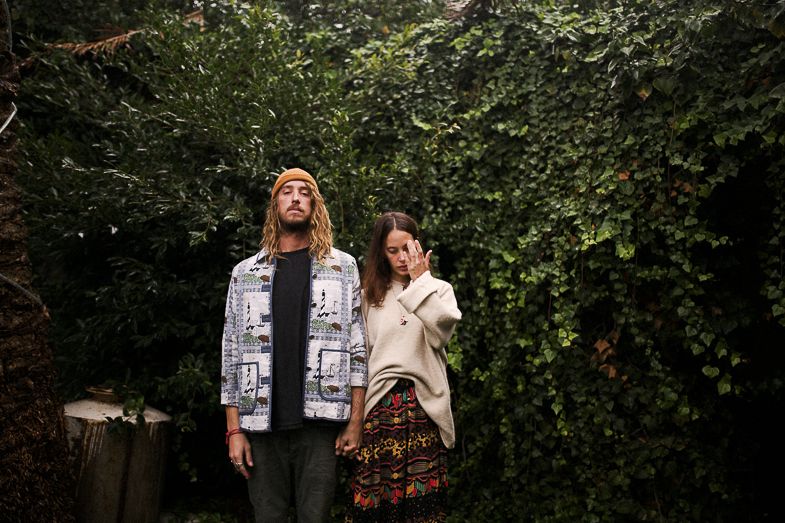A romantic couple stands against a lush green hedge backdrop in casual fall attire.