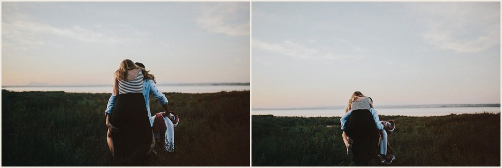 A silhouetted couple standing in a grassy field at sunset with water in the background.