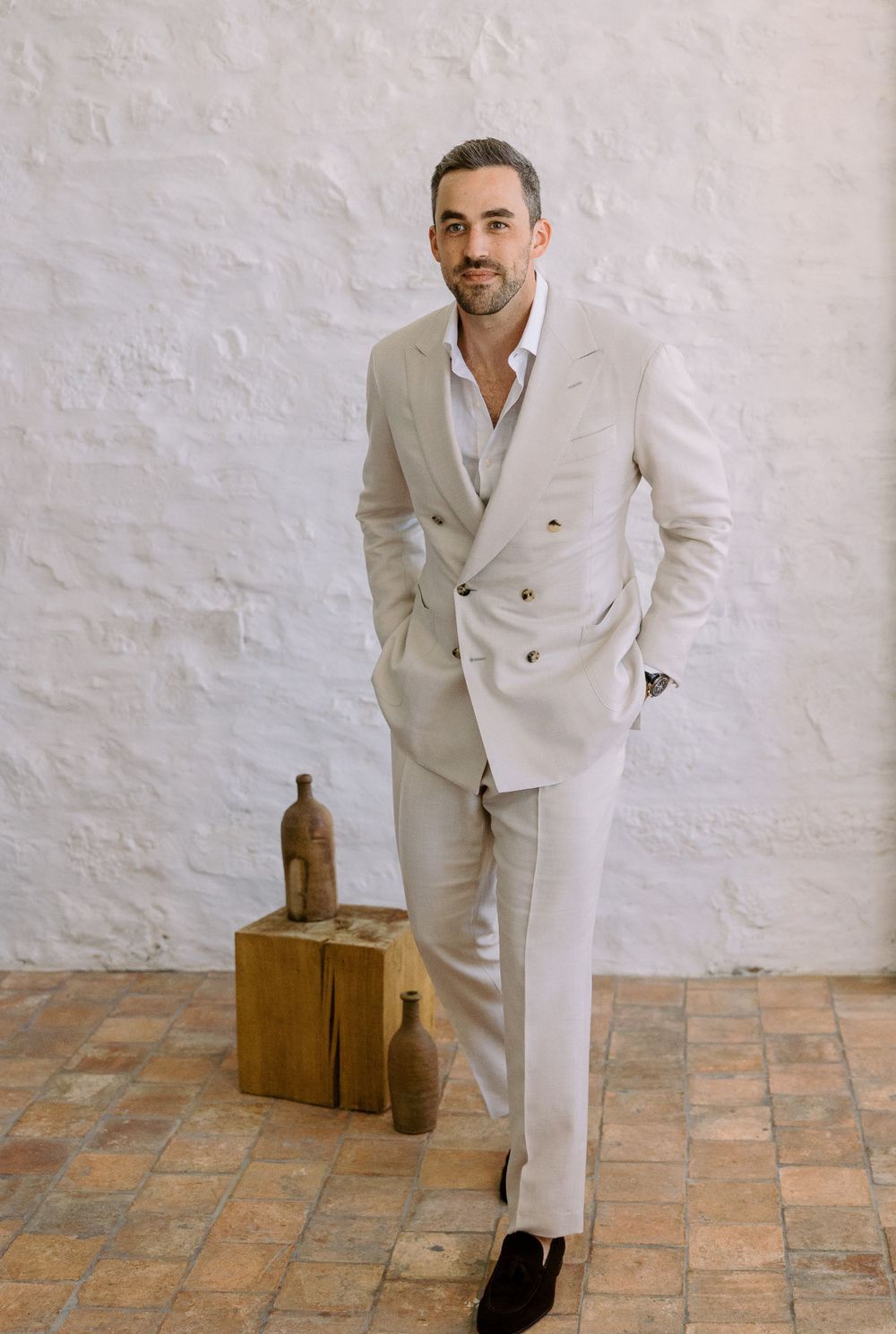 A stylish groom in a cream-colored suit poses against a white textured wall.