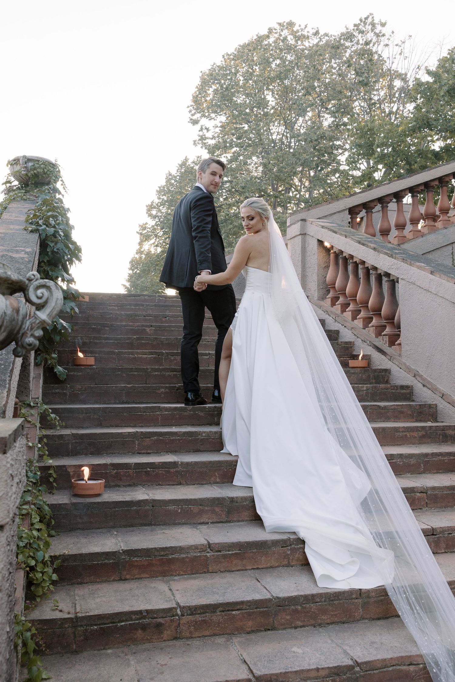 Wedding couple ascending ornate stone stairs decorated with candles and greenery.