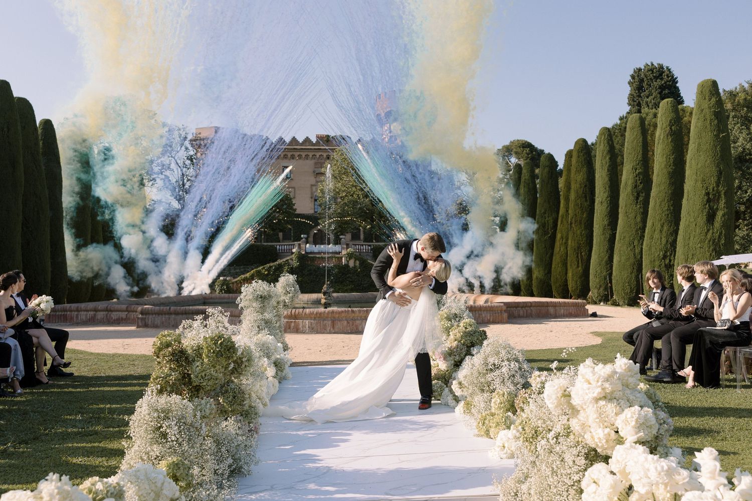 Wedding ceremony with smoke effects and white floral decorations along outdoor aisle.