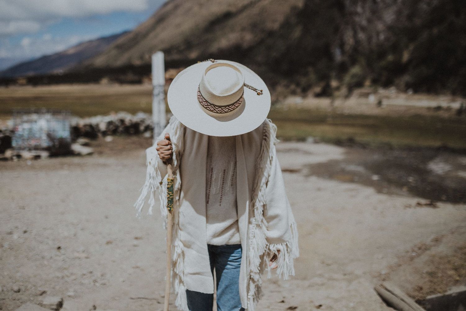Figure in white sweater and wide brim hat stands against mountainous landscape backdrop.