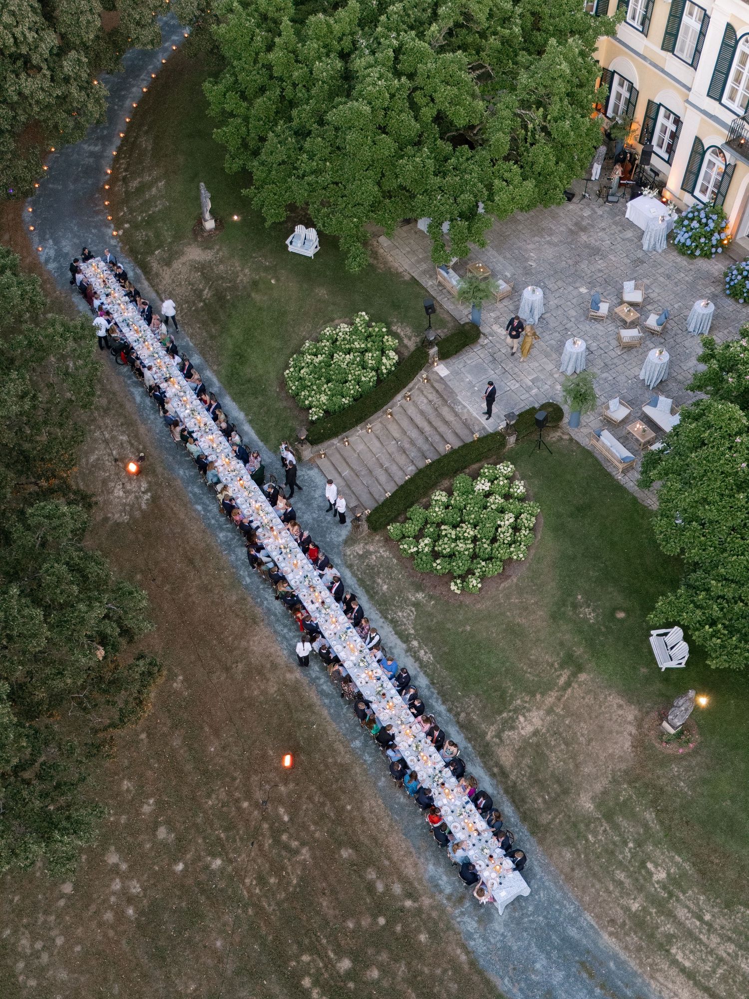 Aerial view of a long outdoor wedding reception dinner table set in a garden estate at dusk.