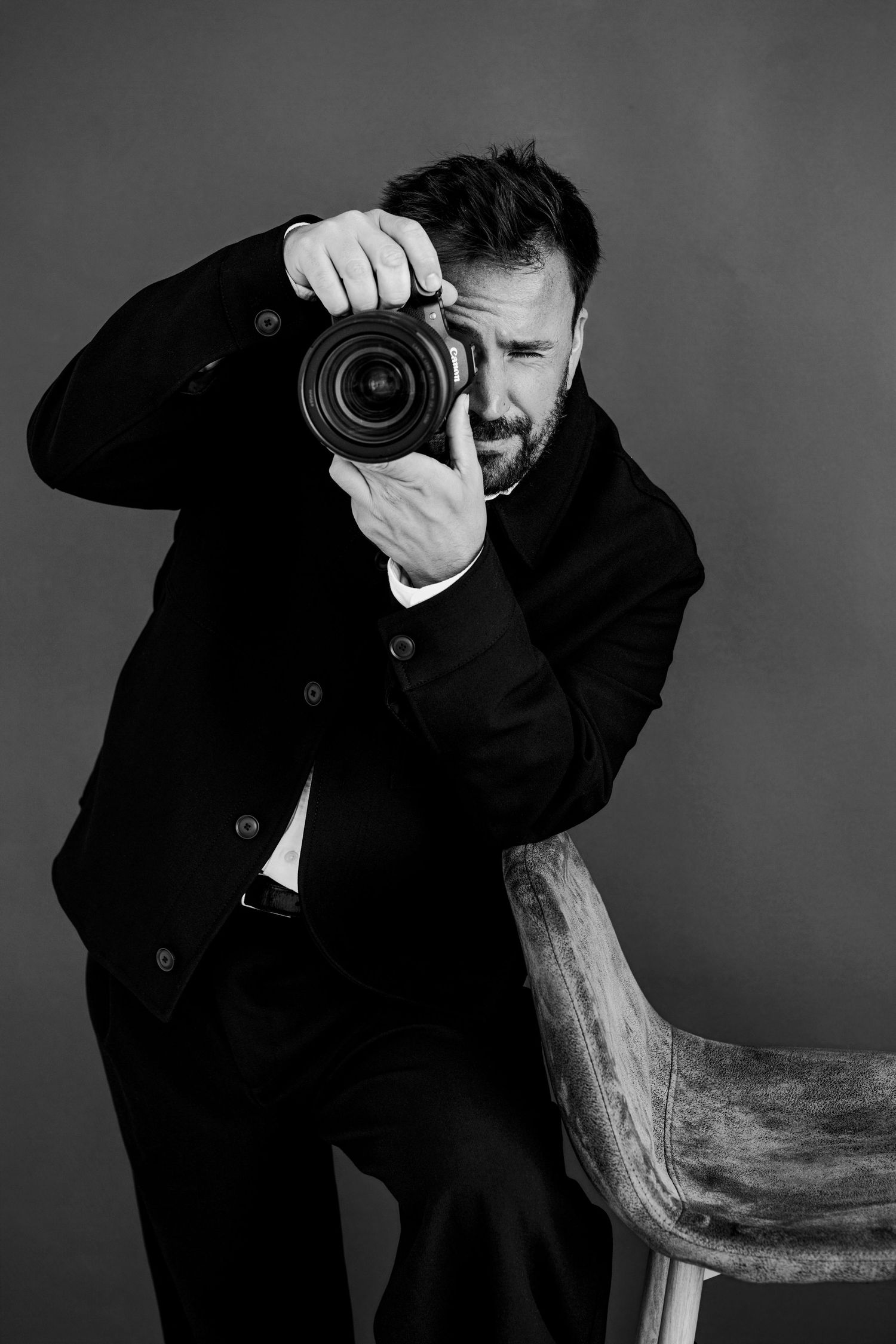 Black and white portrait of a bearded man in a suit holding a DSLR camera up to his eye while leaning on a chair.